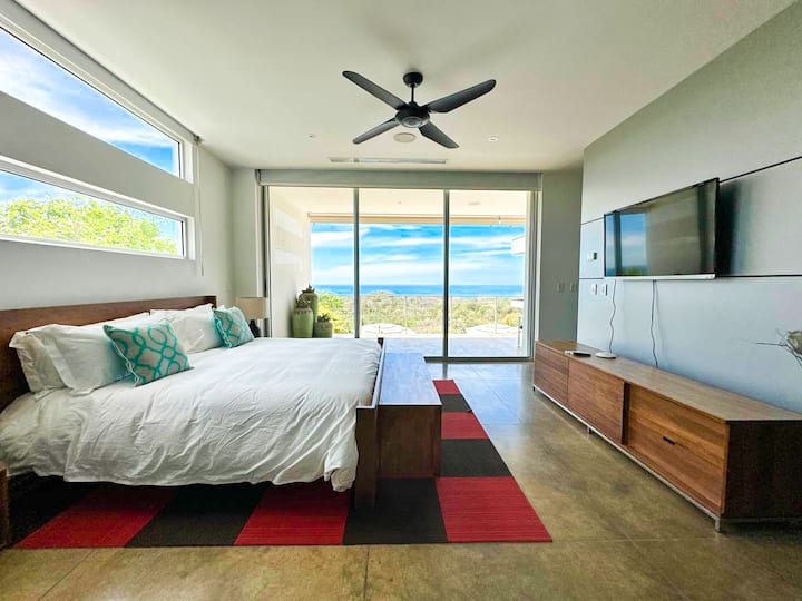 - Impresionante dormitorio con ventanas del piso al techo que muestran vistas panorámicas del aire libre.
- Diseño moderno con TV inteligente y muebles elegantes, incluida una gran cama con marco de madera.
- Una alfombra colorida agrega un contraste audaz a los tonos neutros del espacio.