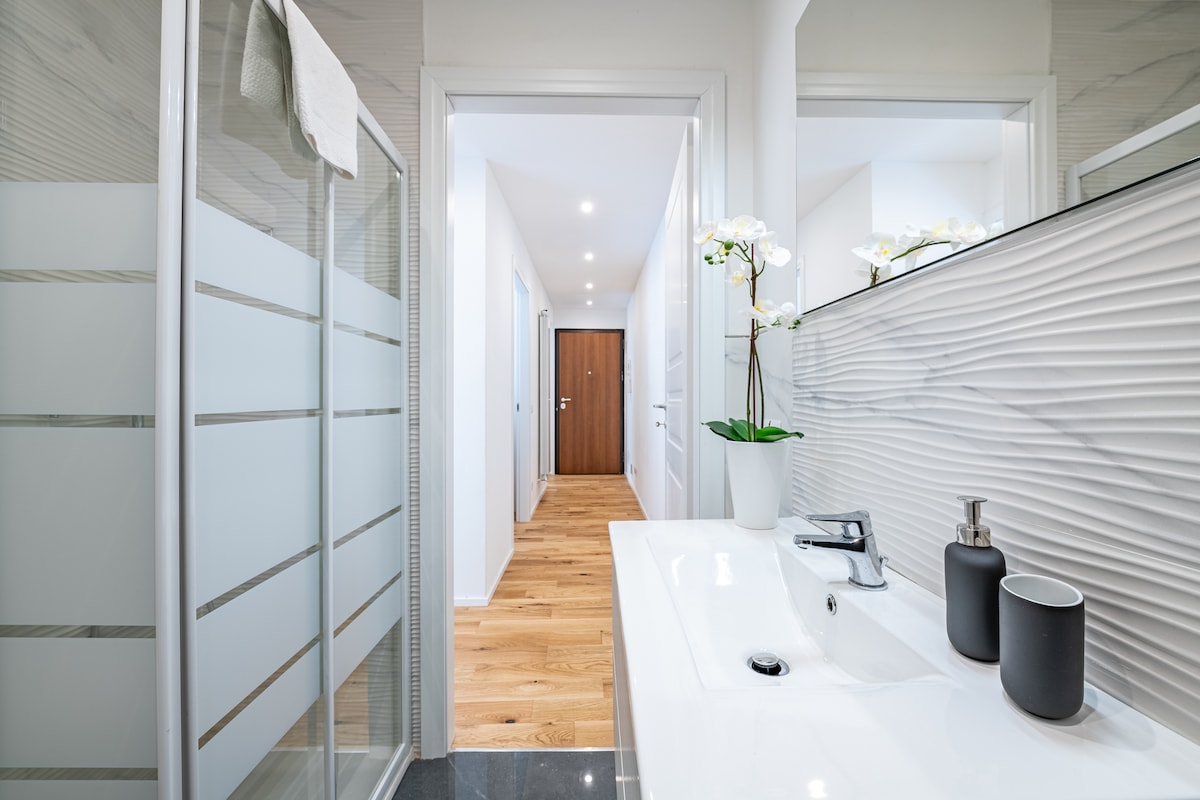 A modern bathroom features a spacious shower with glass doors, complemented by a sleek sink area. Decorative elements include potted orchids and minimalist toiletries, while the hallway is illuminated by recessed lighting, enhancing the overall brightness of the space.