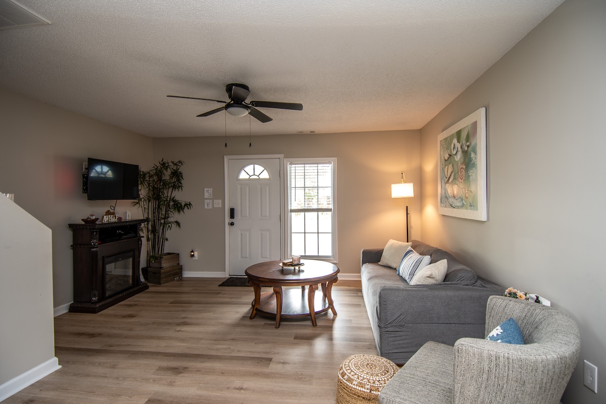 The living area presents an open layout with a comfortable gray sofa positioned near a wooden coffee table. A ceiling fan is installed above, while a small decorative chair adds to the seating options. Natural light enters through the front door and window, creating a bright space.
