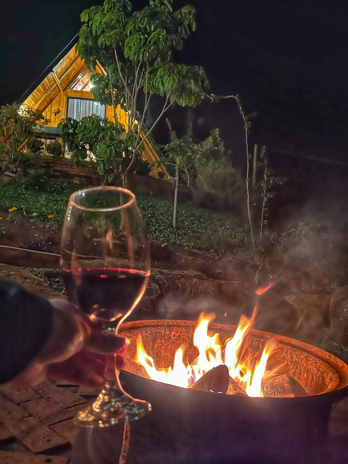 A fire pit is surrounded by warm flames, casting a soft glow in the evening. In the foreground, a hand holds a glass of red wine. The cabin, softly illuminated, is visible in the background, nestled among greenery.