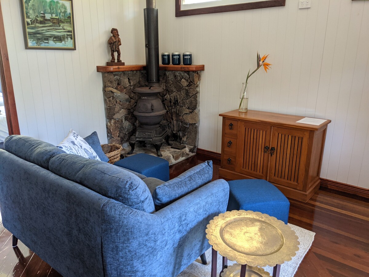 A cozy living area features a blue sofa and two matching ottomans arranged around a stone fireplace. A wooden cabinet stands against the wall, complemented by a decorative golden tray on the floor. Natural light filters in, enhancing the inviting space.