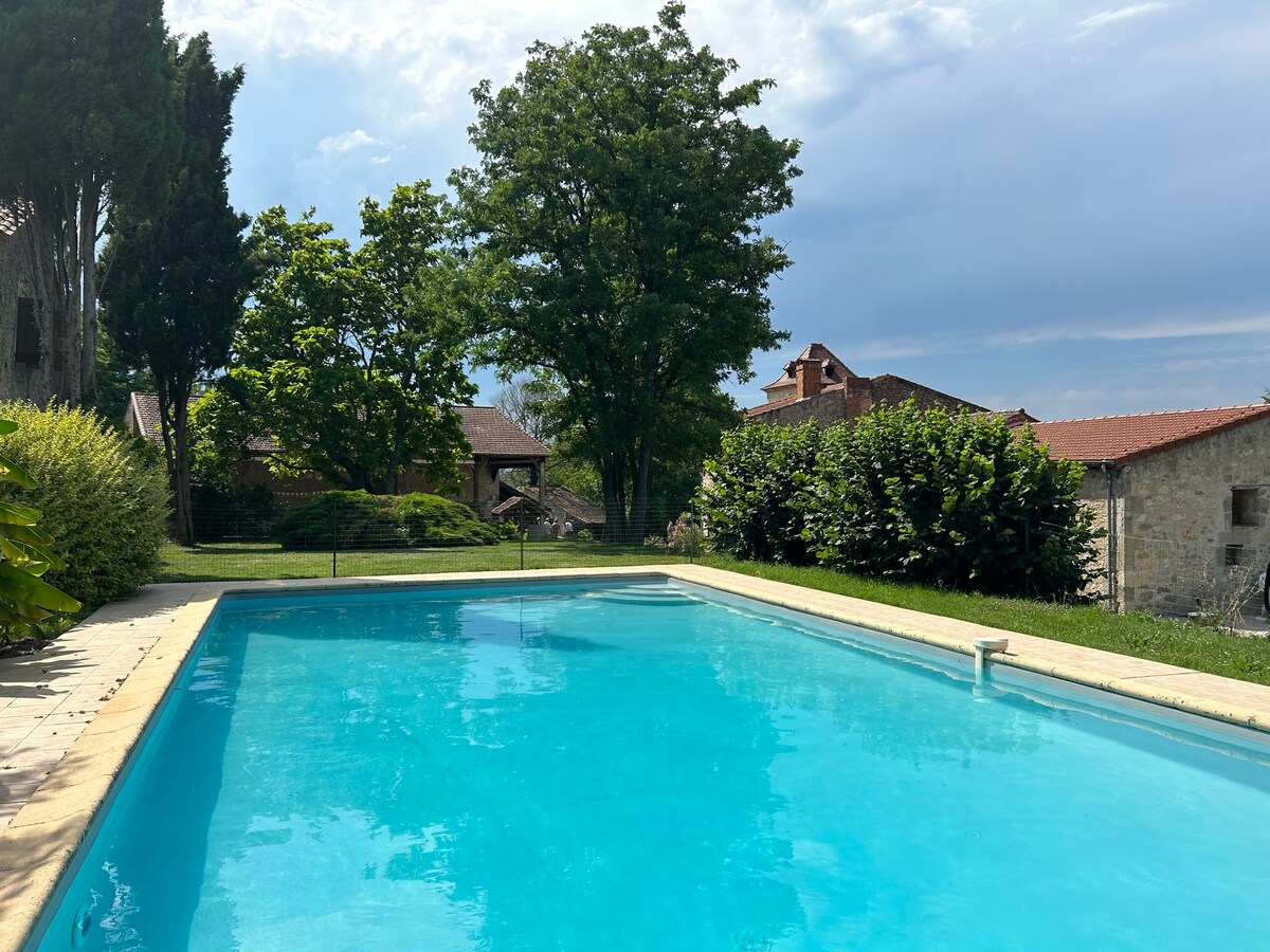 A tranquil pool area is highlighted, featuring a clear blue swimming pool surrounded by lush greenery. Tall trees provide shade, and the distant buildings present a rustic charm, enhancing the peaceful atmosphere of the outdoor space.