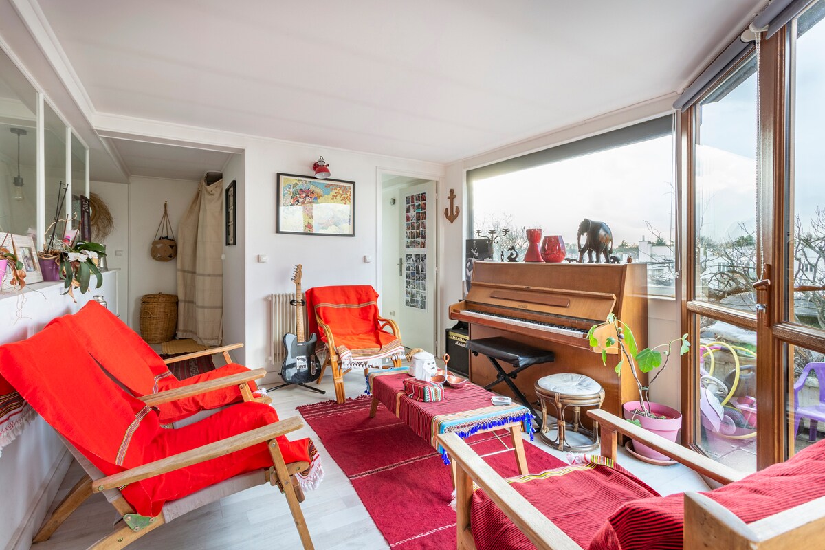 A bright living space features comfortable seating with red cushions, complemented by a wooden piano and eclectic decor. Large glass windows allow natural light to fill the room, offering views of the surroundings. Decorative plants and artwork add character to this inviting area.
