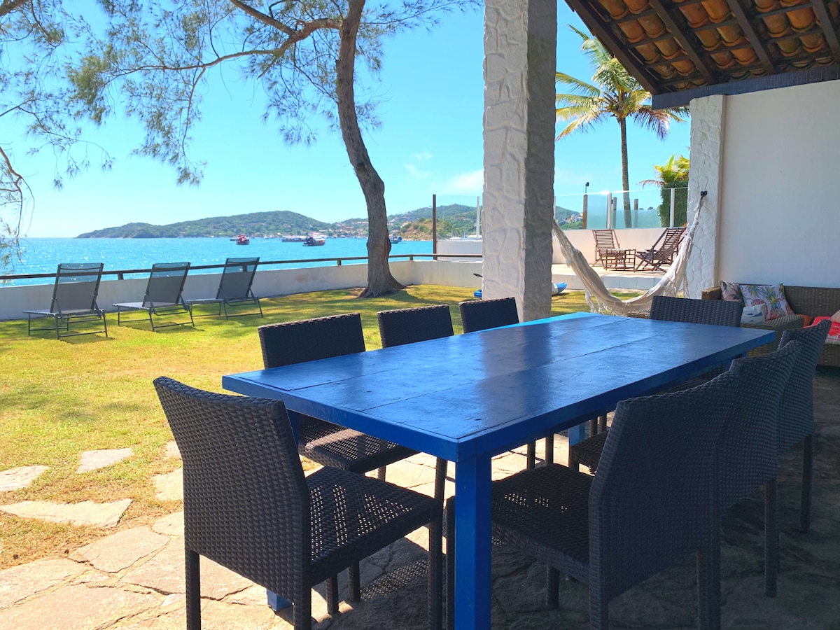 An outdoor dining area features a large blue table surrounded by eight dark chairs. A lush green lawn extends towards the coastline, where the sea is visible under clear blue skies. Nearby, lounge chairs are arranged for relaxation.