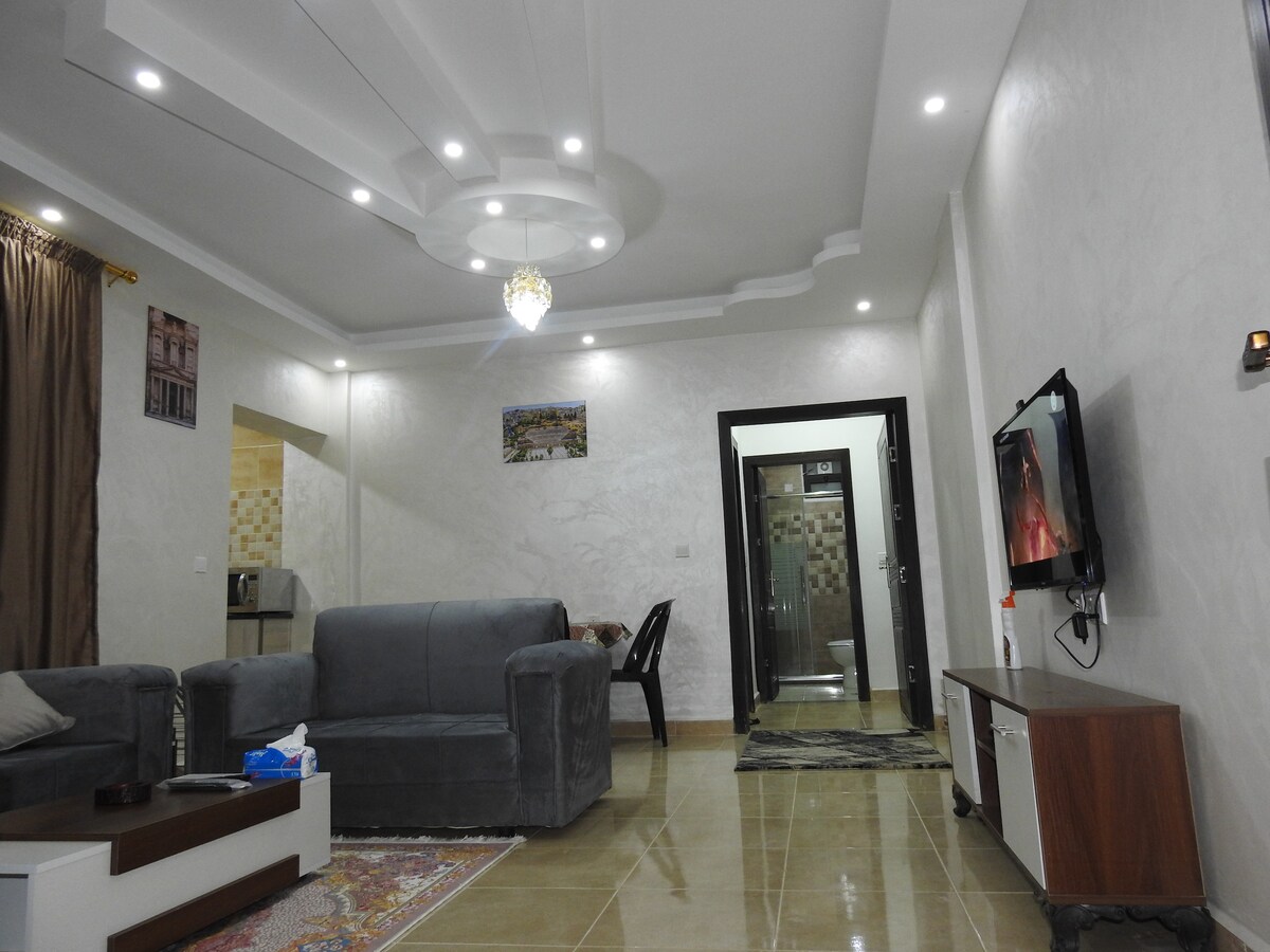 A spacious living area is showcased with a soft gray sofa facing a wall-mounted television. Ambient lighting is provided by recessed ceiling fixtures and a chandelier. A small coffee table sits on a patterned rug, with a glimpse of a kitchen area and a bathroom visible in the background.