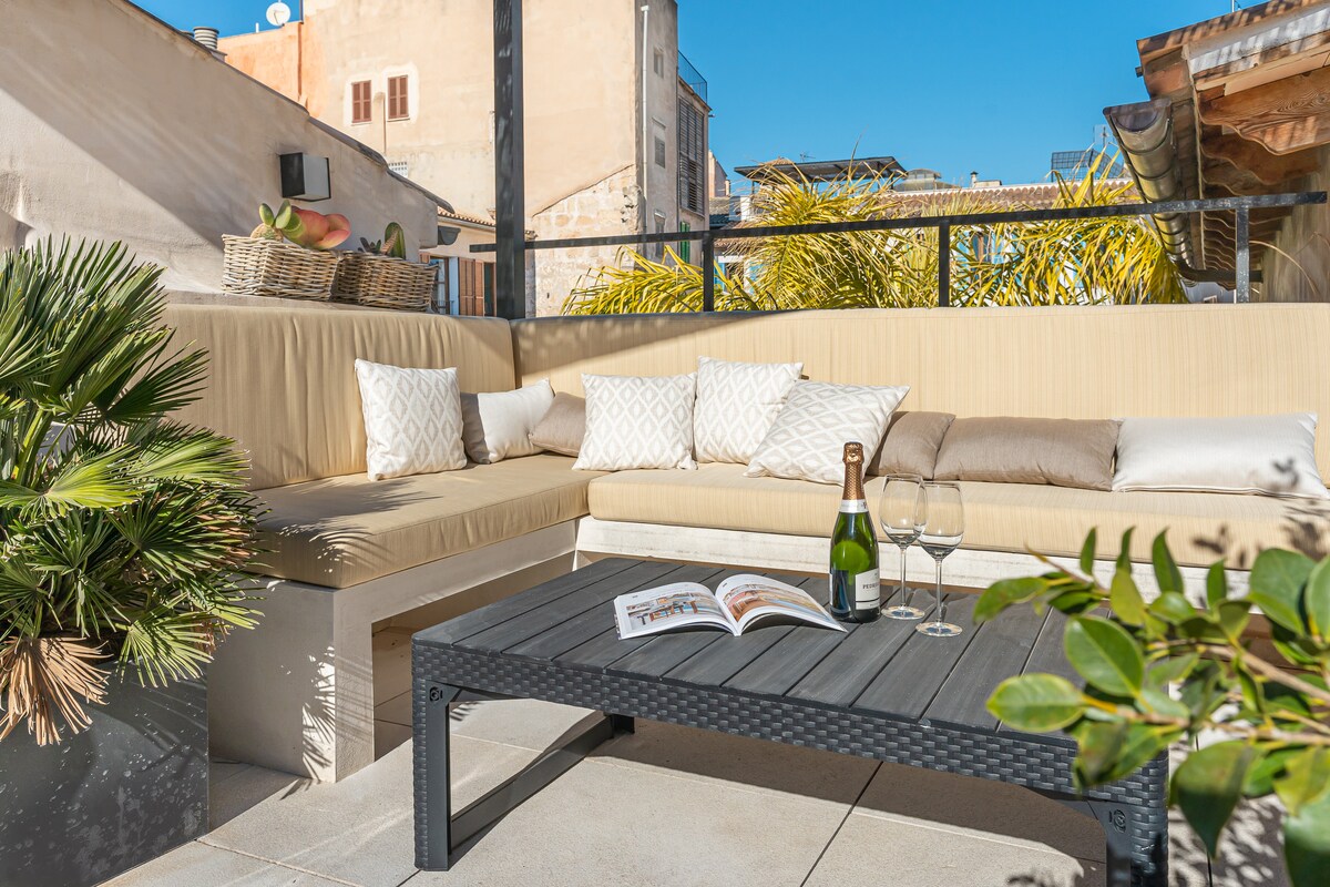An outdoor lounge area features a sectional sofa with neutral cushions and a low coffee table. A bottle of champagne and two glasses rest on the table beside a magazine. Lush greenery adds a touch of nature to the space, which is bathed in sunlight.