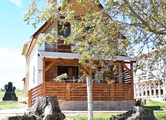 The charming house is surrounded by a lush green lawn and features a welcoming wooden porch. Natural light highlights the exterior details, including wooden accents and large windows. A fence encloses the property, providing an added sense of privacy and tranquility.