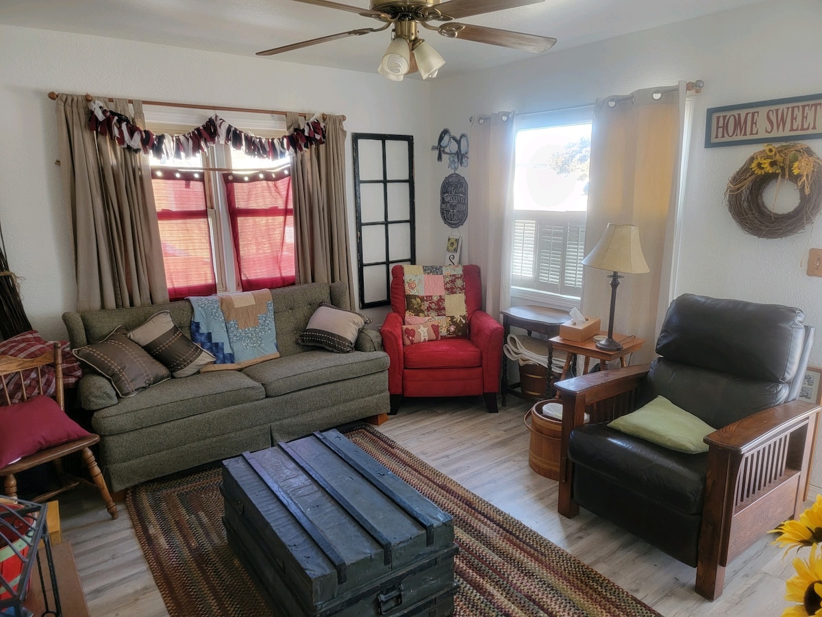 A cozy living room features a soft gray sofa and a red accent chair, complemented by a comfortable recliner. A wooden coffee table rests on a textured area rug, while large windows allow natural light to fill the space, accentuated by warm curtains and decorative wall art.