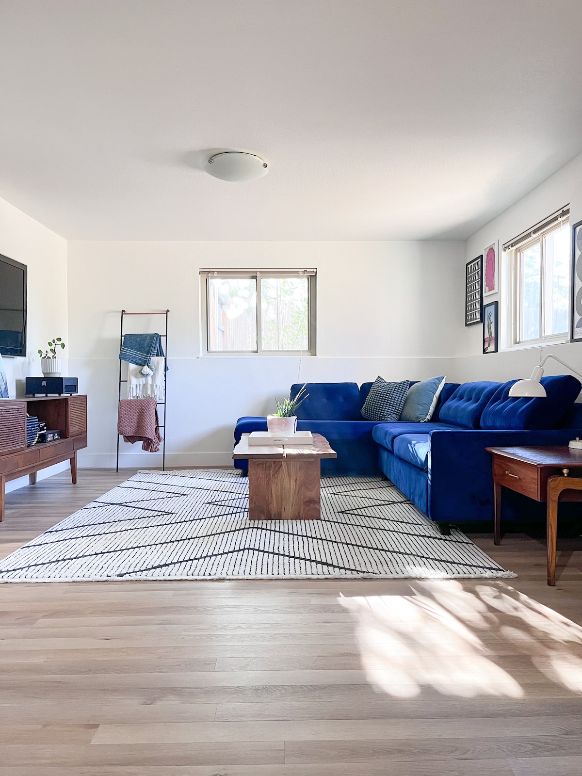 The living area features a deep blue sectional sofa, accompanied by a wooden coffee table at its center. Natural light filters through two windows, illuminating the light wood flooring. A patterned area rug adds texture, with a small side table placed next to the sofa, highlighting a welcoming atmosphere.