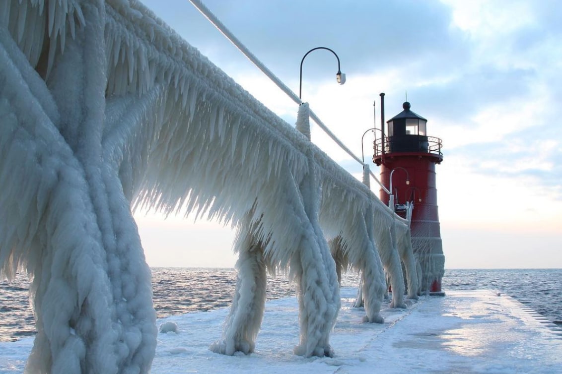 A frozen pier extends into the lake, adorned with ice formations that create a glistening texture. The red lighthouse stands at the end, contrasting against the icy structures and soft blue hues of the water and sky.