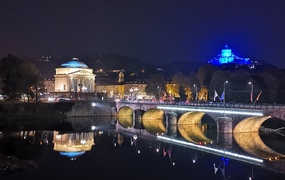 A scenic nighttime view of the iconic Mole Antonelliana and the nearby hilltop church, beautifully illuminated in blue light. The Po River reflects the architectural details of the bridge and the historical buildings along the riverbank, creating a serene atmosphere.