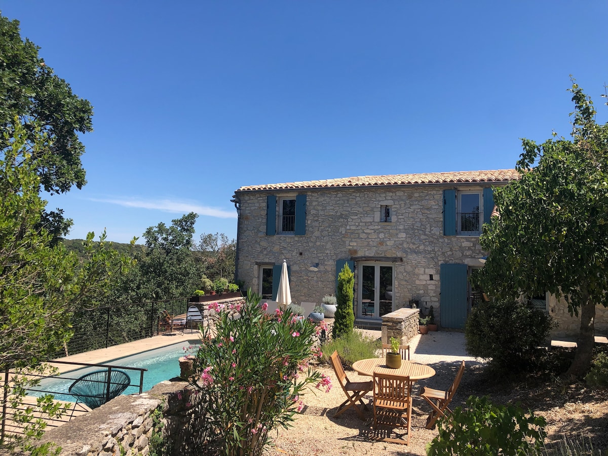 A charming stone house features blue shutters and a spacious terrace. A private pool, surrounded by greenery and flowers, can be seen in the foreground. Outdoor dining furniture is arranged nearby, inviting relaxation in the natural setting.
