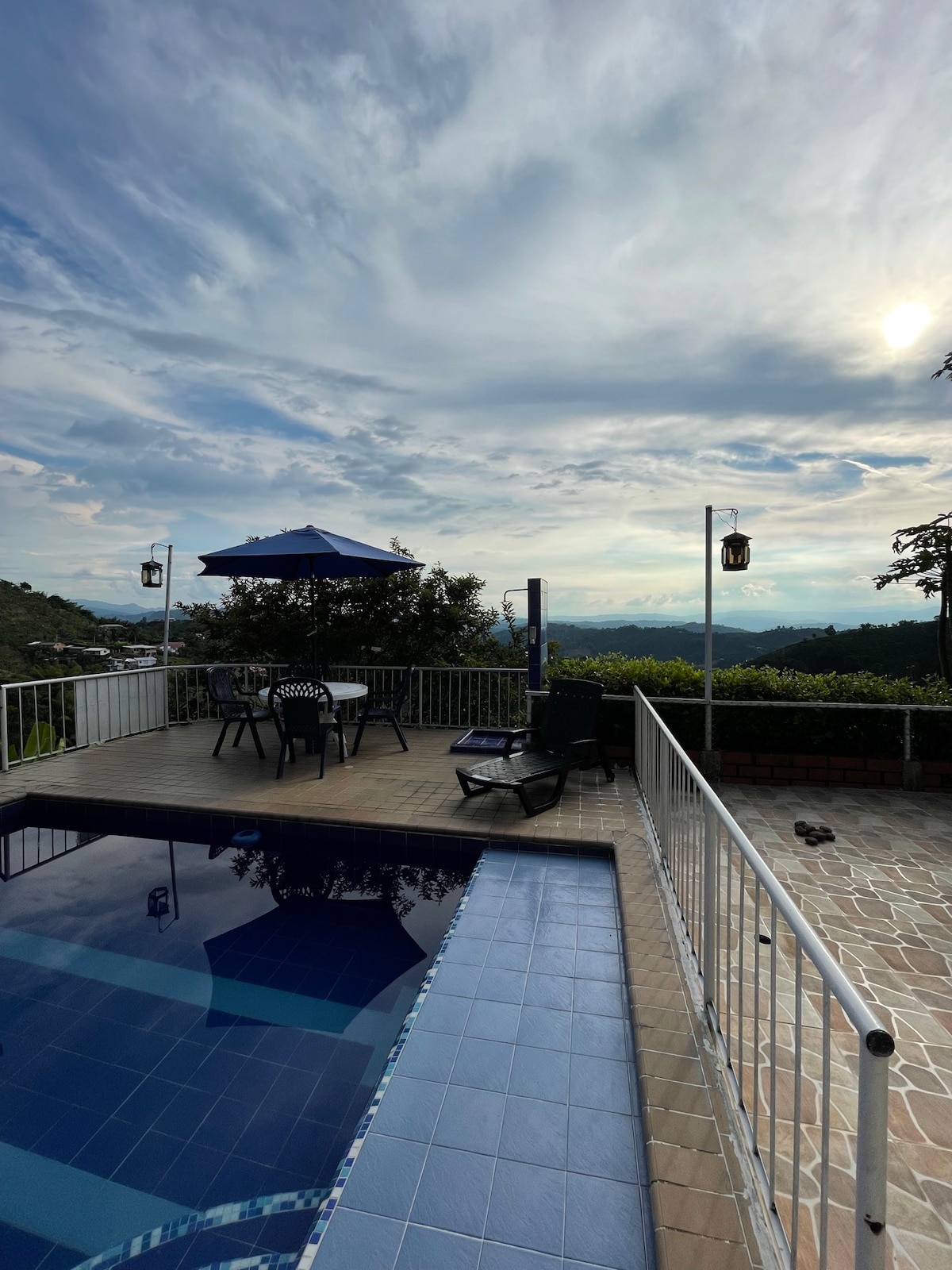 An outdoor space showcases a swimming pool with a tiled edge, surrounded by a white railing. A patio with a table, chairs, and an umbrella overlooks a panoramic view, with lanterns hanging nearby. The sky is filled with soft clouds and gentle light.