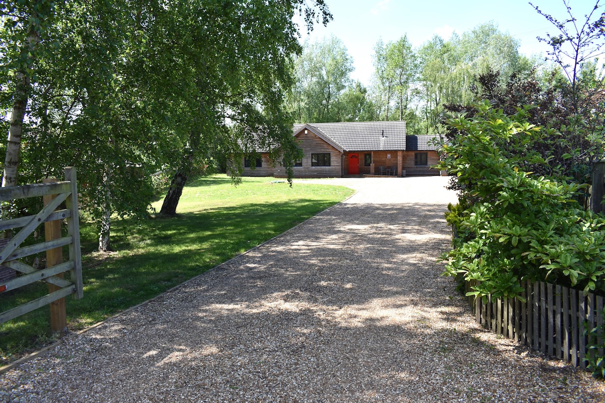 A gravel driveway leads towards a single-story lodge, framed by lush greenery and trees. The entrance features a striking red door, while well-maintained lawns surround the property, offering a serene and inviting approach to the home.