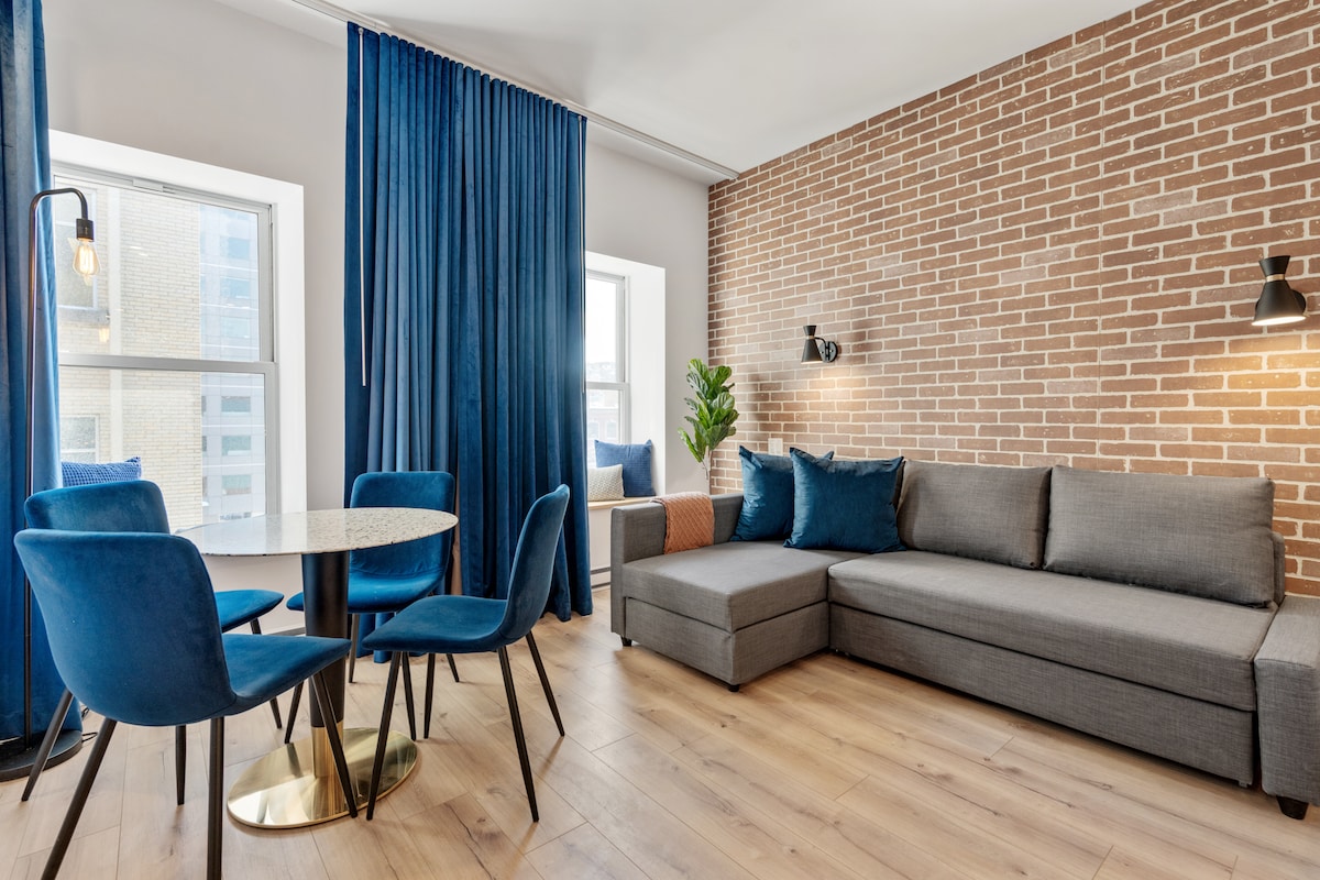 A contemporary living area is showcased, featuring a gray sectional sofa, a round dining table with four blue chairs, and a decorative plant. Large windows are adorned with blue curtains, and exposed brick walls add texture to the space. Soft lighting is provided by wall sconces.