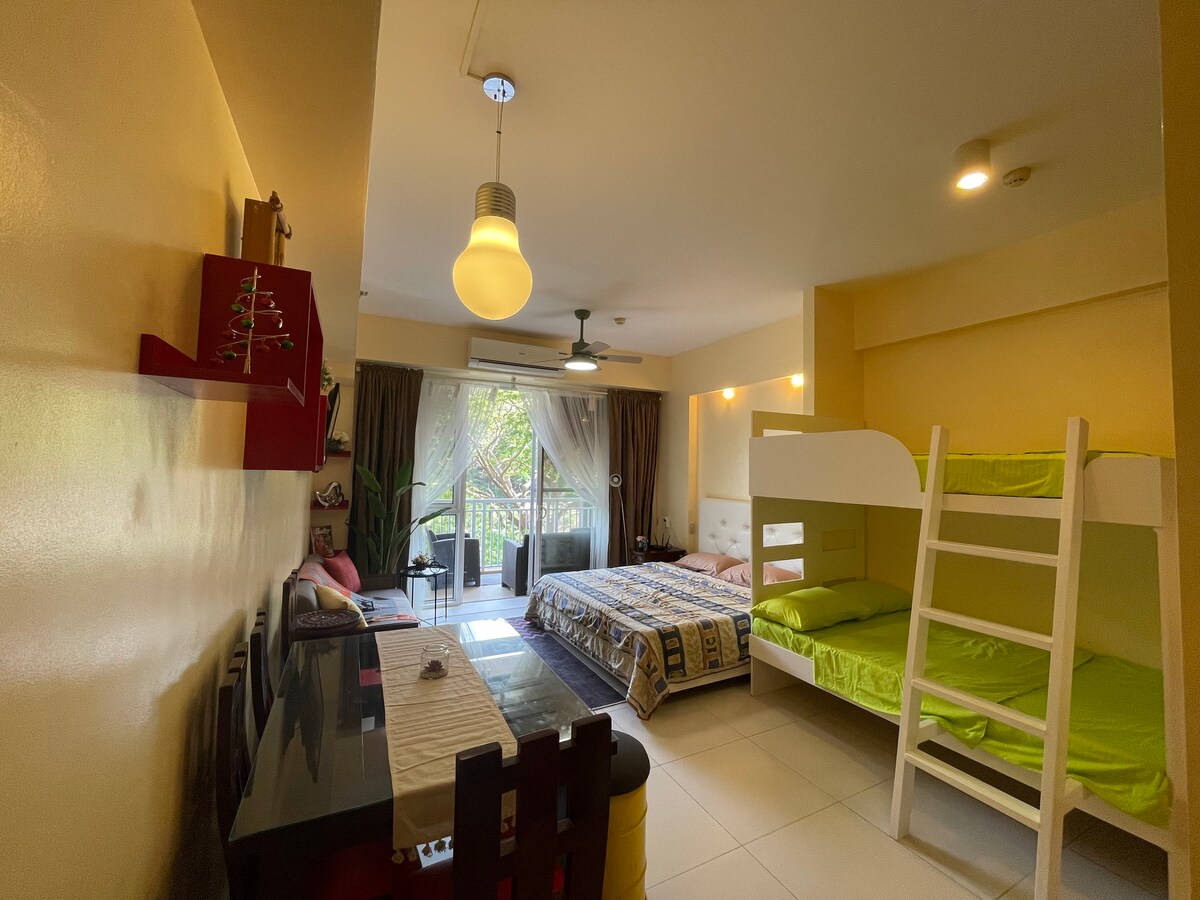 A bright and airy room features a king-size bed and a double deck bed, accommodating up to six guests. A dining area is visible alongside a kitchenette. Large windows provide natural light and access to a terrace with a view, enhancing the spaciousness of the room.