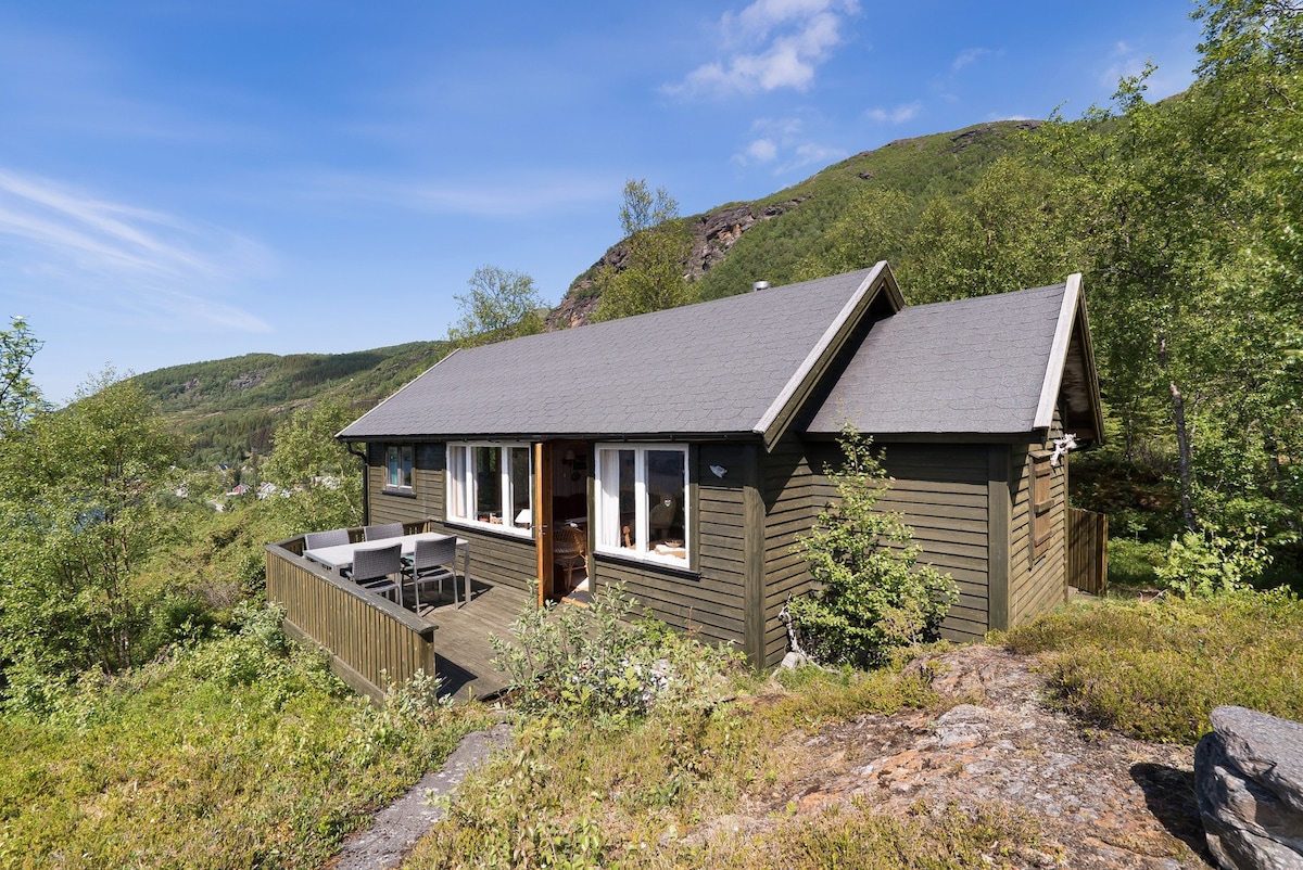 A charming wooden cabin is surrounded by greenery, featuring a deck with outdoor seating. Large windows allow natural light to fill the interior, and scenic mountain views can be seen in the background, enhancing the tranquil setting.