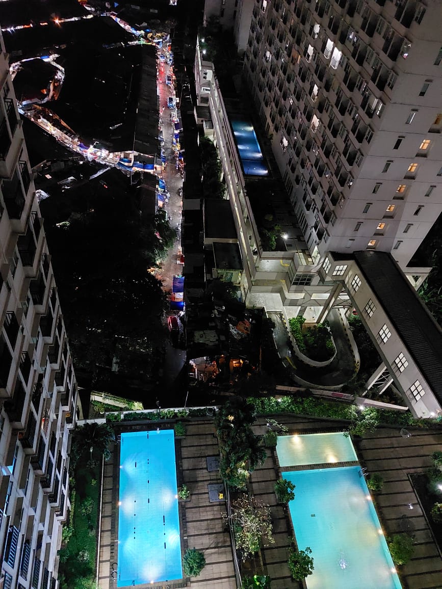 An aerial view reveals two swimming pools surrounded by green landscaping, illuminated softly at night. Nearby, a lighted pathway and structures hint at additional amenities in the bustling area, while vibrant market stalls are visible in the distance.