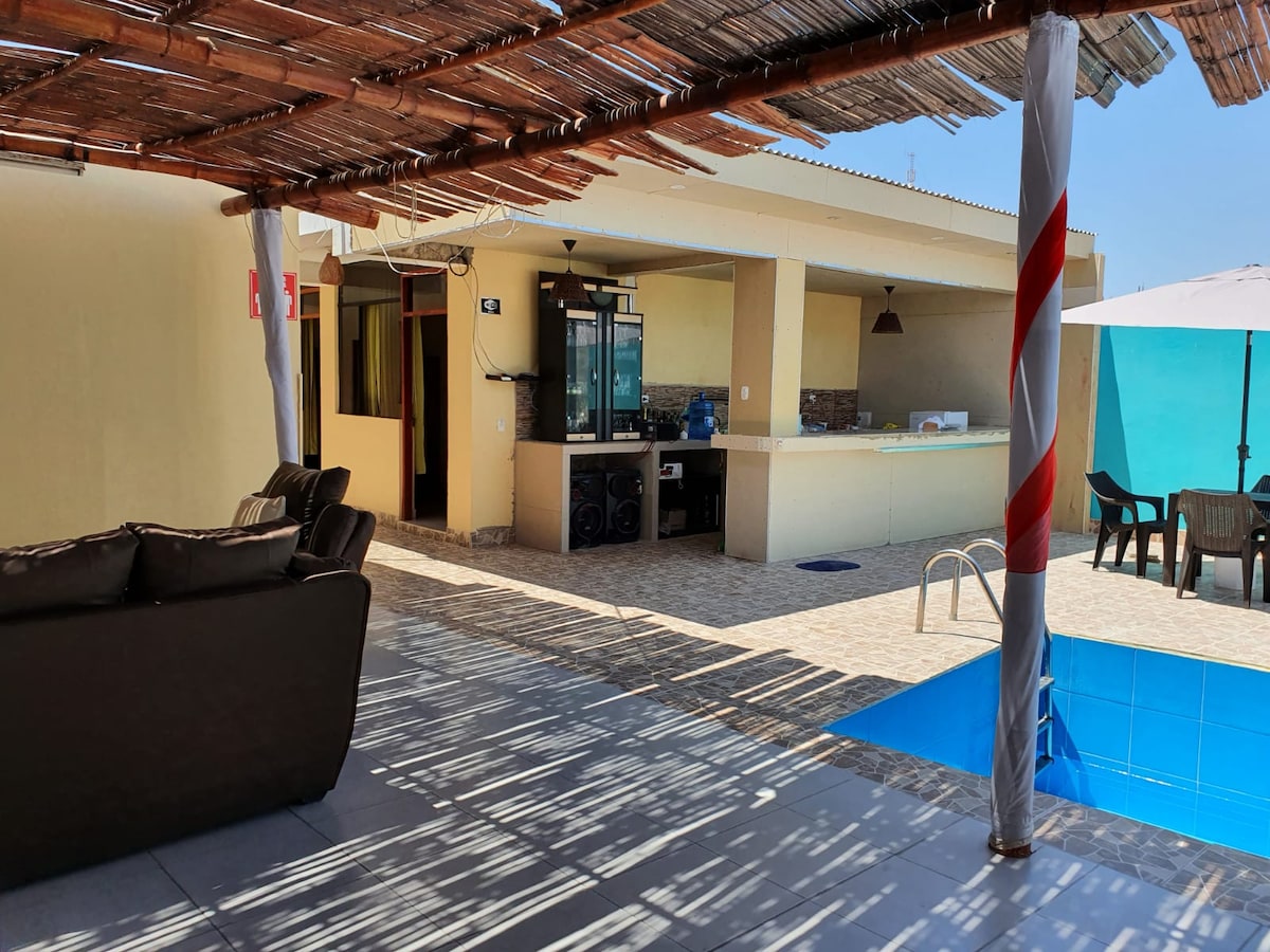 A shaded outdoor seating area features comfortable sofas, providing a relaxed space near a shimmering pool. A bar area is visible in the background, complete with counter space and appliances. Bright blue walls enhance the open atmosphere, and an umbrella offers additional shade.