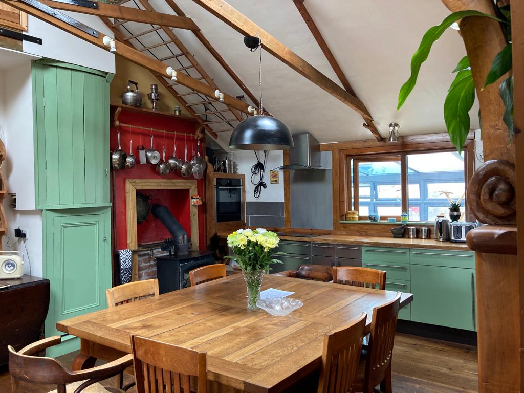 An open-plan kitchen features wooden beams and high ceilings, enhancing the light-filled space. A wooden dining table with eight chairs is set at the center. Green cabinets complement the rustic decor, while a cozy wood burner is visible in the background.
