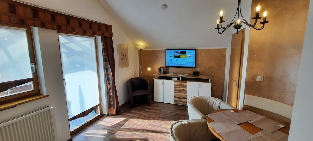 A cozy living area is featured, accented by wood-toned flooring. A flat-screen TV is mounted on a stylish cabinet, while a comfortable armchair and two light-colored chairs are positioned nearby. Large windows invite natural light, and a chandelier adds a touch of warmth.