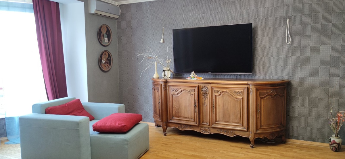 A spacious living area features a light blue couch with red accent pillows, positioned opposite a wooden sideboard holding decorative items. A large flat-screen TV is mounted on the wall, while bright natural light enters through a window covered with red curtains.