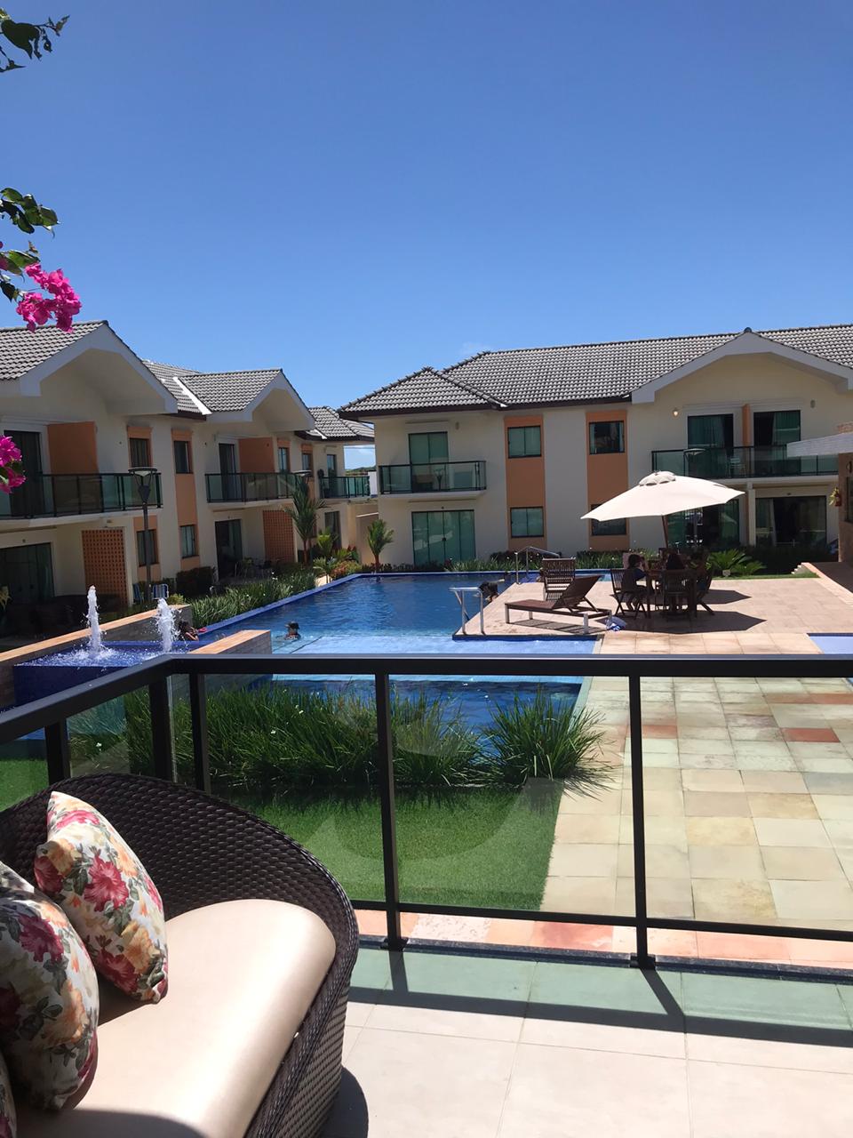 A serene view showcases a well-maintained pool surrounded by lounge chairs and umbrellas. Lush greenery and vibrant flowers complement the outdoor space, while modern buildings frame the scene, reflecting the clear blue sky overhead.