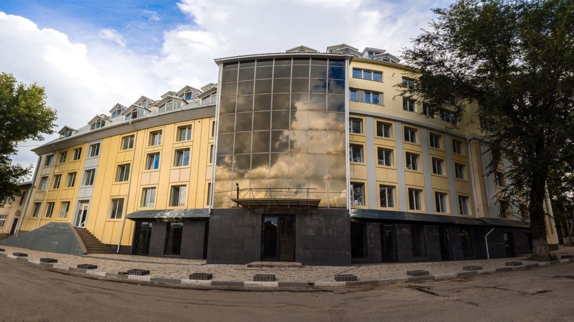 The building features a modern façade with large glass panels reflecting the sky, complemented by a smooth stone base. The structure has multiple levels with a series of windows, providing natural light to the interior. Surrounding trees and a paved walkway enhance the urban landscape.