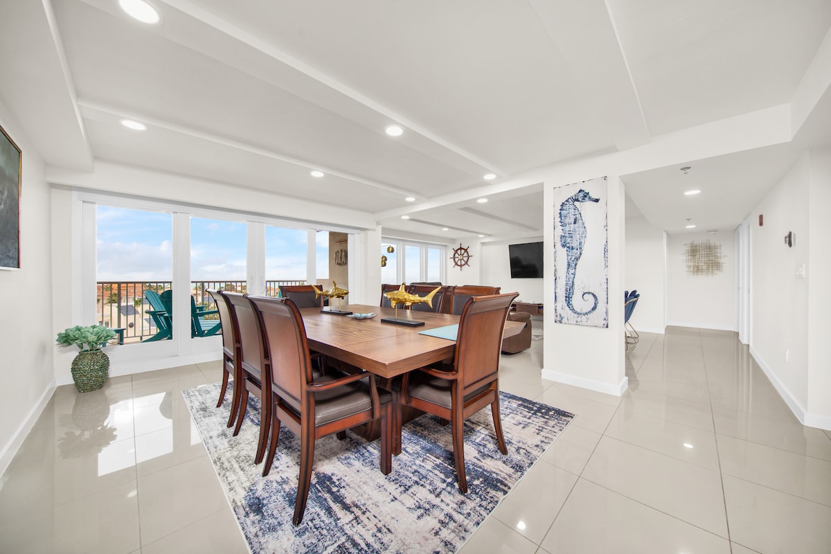A spacious dining area features a wooden table surrounded by eight chairs, positioned on a decorative rug. Large windows provide ample natural light and offer a beach view. The room is complemented by a peaceful color palette and modern decor, enhancing the inviting atmosphere.