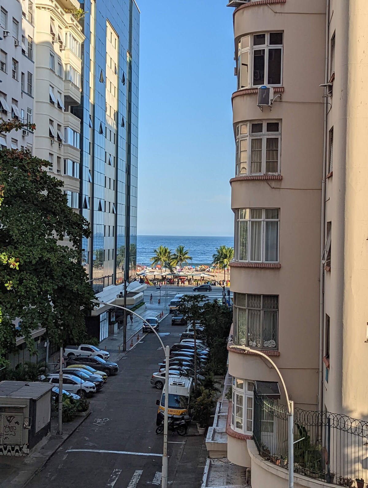 A view is presented from a window overlooking a vibrant street leading to the beach. Palm trees are visible near the shoreline, and the blue ocean can be seen in the distance. The architecture includes a mix of tall buildings and quaint structures.