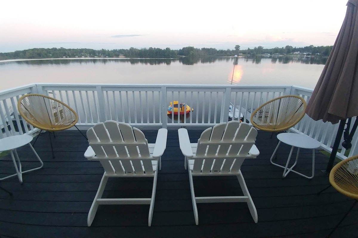 A spacious deck is furnished with two white Adirondack chairs and a small table. The calm waters of Hardwood Lake are visible beyond the railing, reflecting the soft hues of the evening sky. A colorful boat floats gently on the lake's surface.