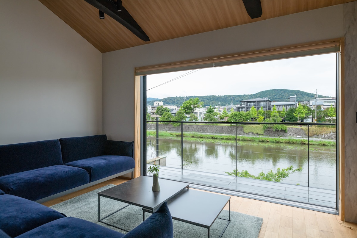 A spacious living area features a deep blue sectional sofa with a sleek coffee table positioned in front. Large sliding glass doors provide a panoramic view of the river and distant hills, creating a sense of openness and connection to the natural surroundings.