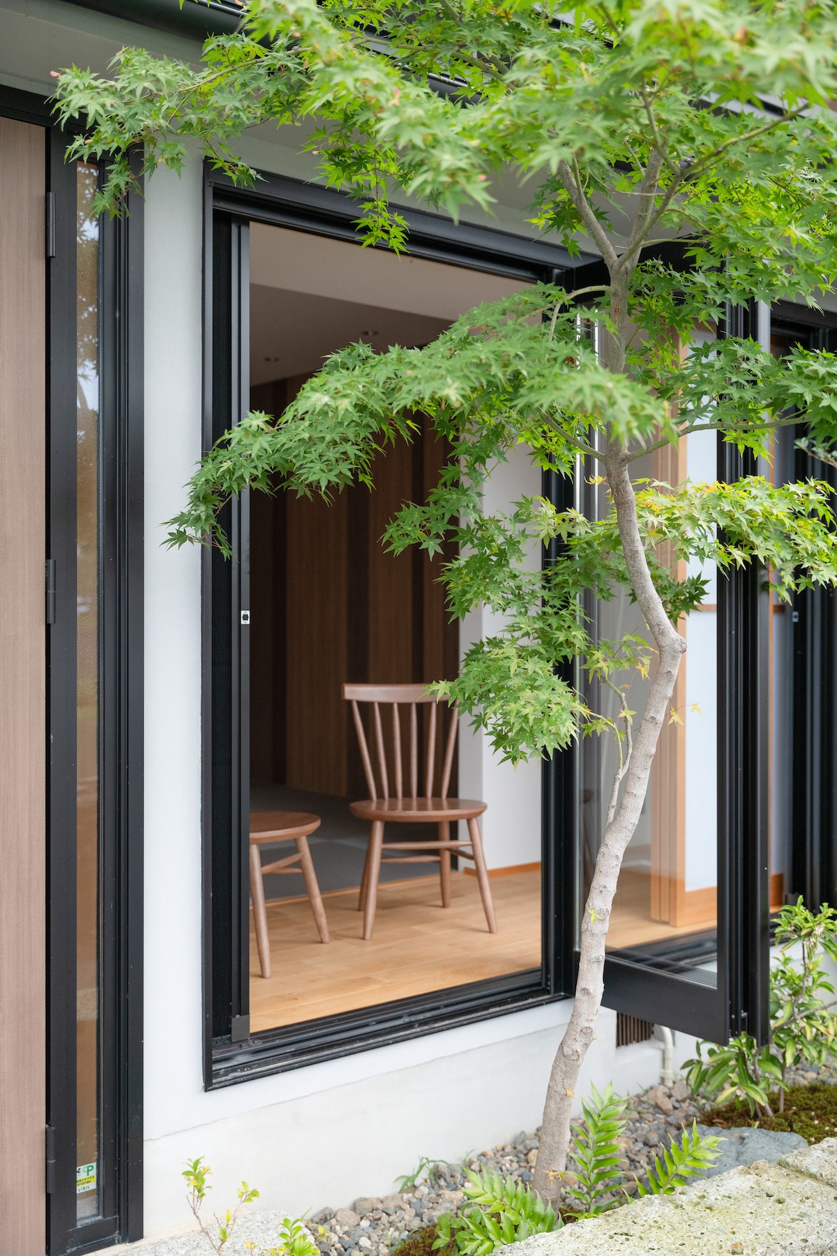 A serene corner of the building is framed by lush greenery. Two wooden chairs are positioned near a partially open window, allowing natural light to enter the space. The warm wooden flooring complements the modern interior design visible beyond.