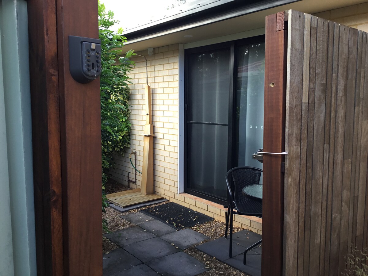 An entrance gate opens to a private courtyard featuring a small outdoor table and chair set on a stone pathway. Lush greenery is visible, and large glass doors lead into the interior space, providing a sense of seclusion and accessibility.