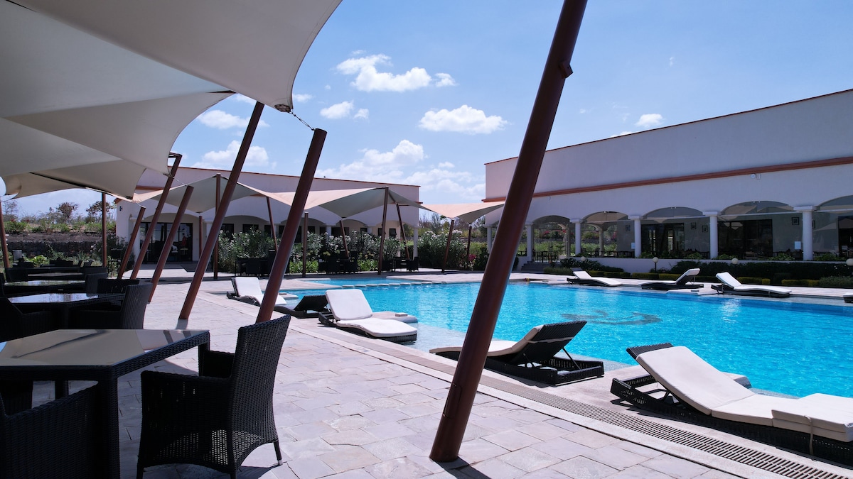 A serene outdoor area features a large swimming pool surrounded by sun loungers and shaded seating. Several cabanas provide cover, with landscaping visible in the background. Clear blue skies and soft clouds create a peaceful atmosphere.