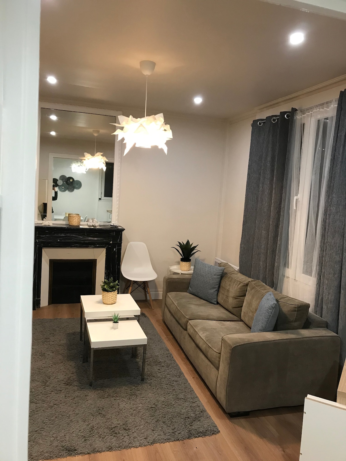 A modern living area is presented with a plush sofa and minimalist coffee table, positioned atop a soft rug. Natural light enters through a large window, framed by dark curtains. A decorative light fixture and potted plants add a touch of warmth.