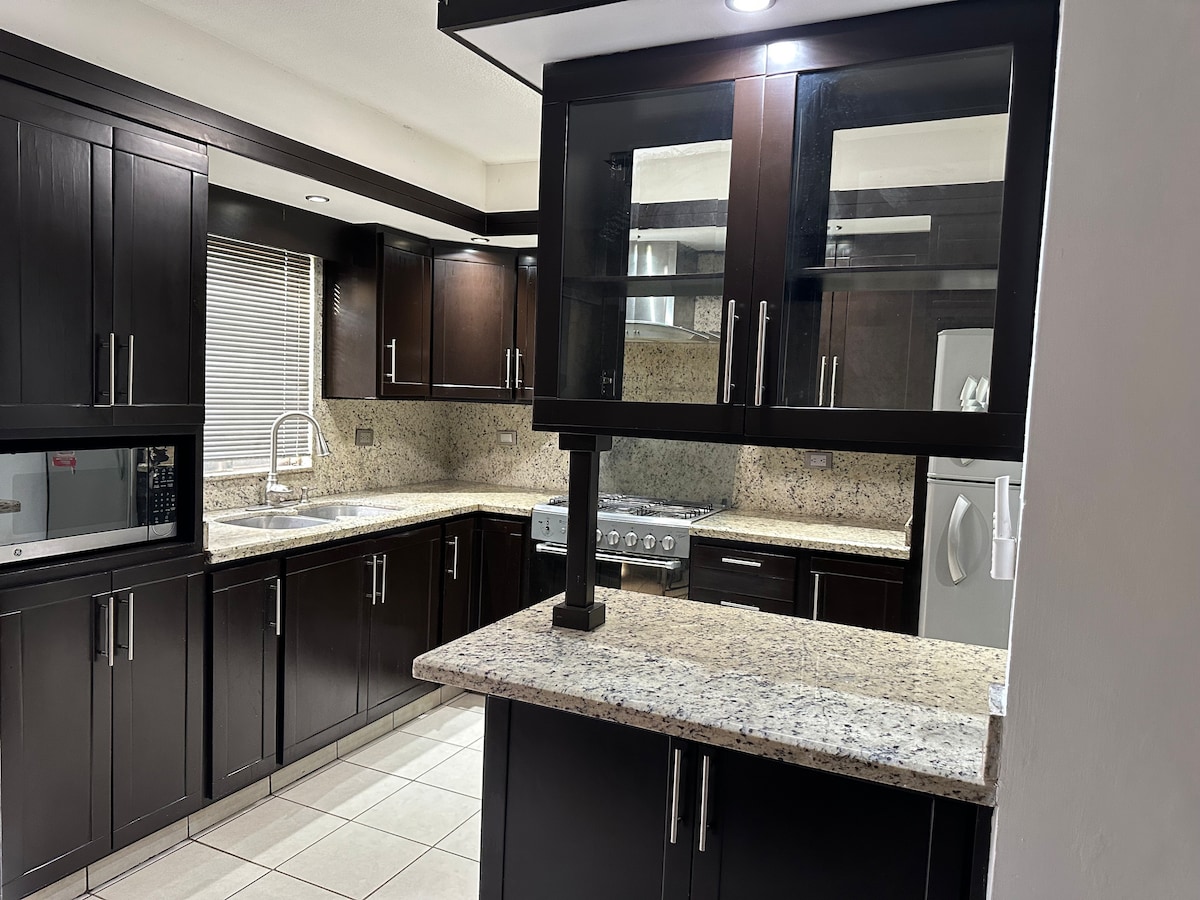The kitchen features dark wood cabinetry complemented by a light granite countertop. A spacious island provides additional prep space, while modern appliances are integrated within the cabinetry. A window allows natural light to illuminate the room, enhancing its functional layout.