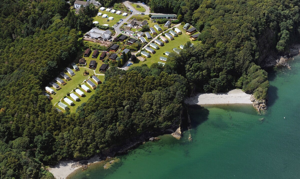 An aerial view captures the peaceful caravan park nestled among lush greenery. The arrangement of holiday homes is visible, surrounded by trees, with a gentle shoreline and clear waters nearby, illustrating a tranquil retreat atmosphere.