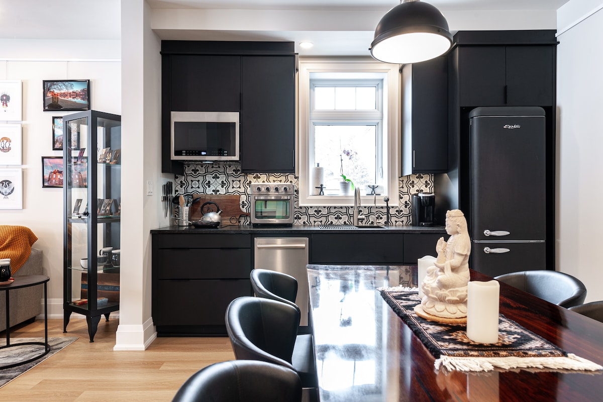 A modern kitchenette and dining area feature sleek black cabinetry and a stylish dining table with black chairs. Decorative ceramic tiles add a decorative touch behind the stainless steel appliances, including a microwave and refrigerator. A sculpture and candles are placed centrally on the table.