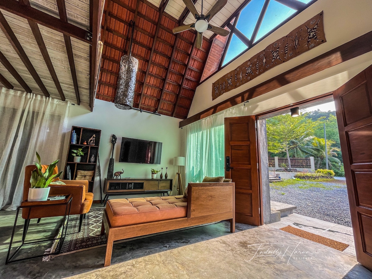 The open living area features high, slanted wooden ceilings with a ceiling fan and large glass doors that allow natural light to fill the space. A comfortable seating area is accompanied by a TV and ornamental plants, providing a welcoming and relaxed atmosphere.