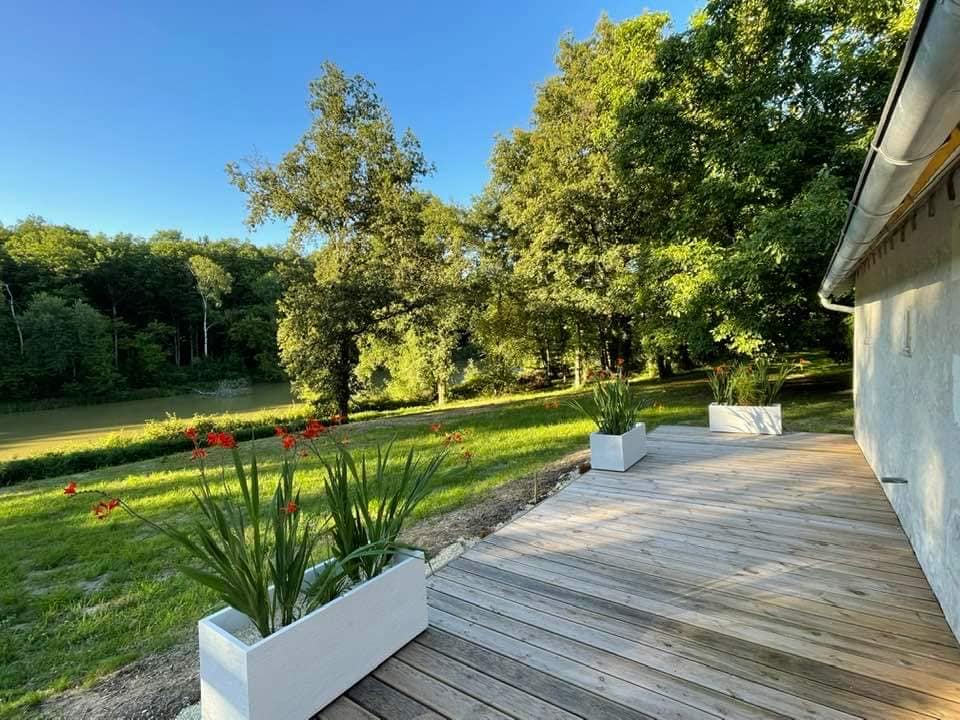 A wooden terrace extends beside a tranquil pond, adorned with greenery. Large planters filled with vibrant flowers offer a pop of color against the serene backdrop of the surrounding trees and lush landscape.