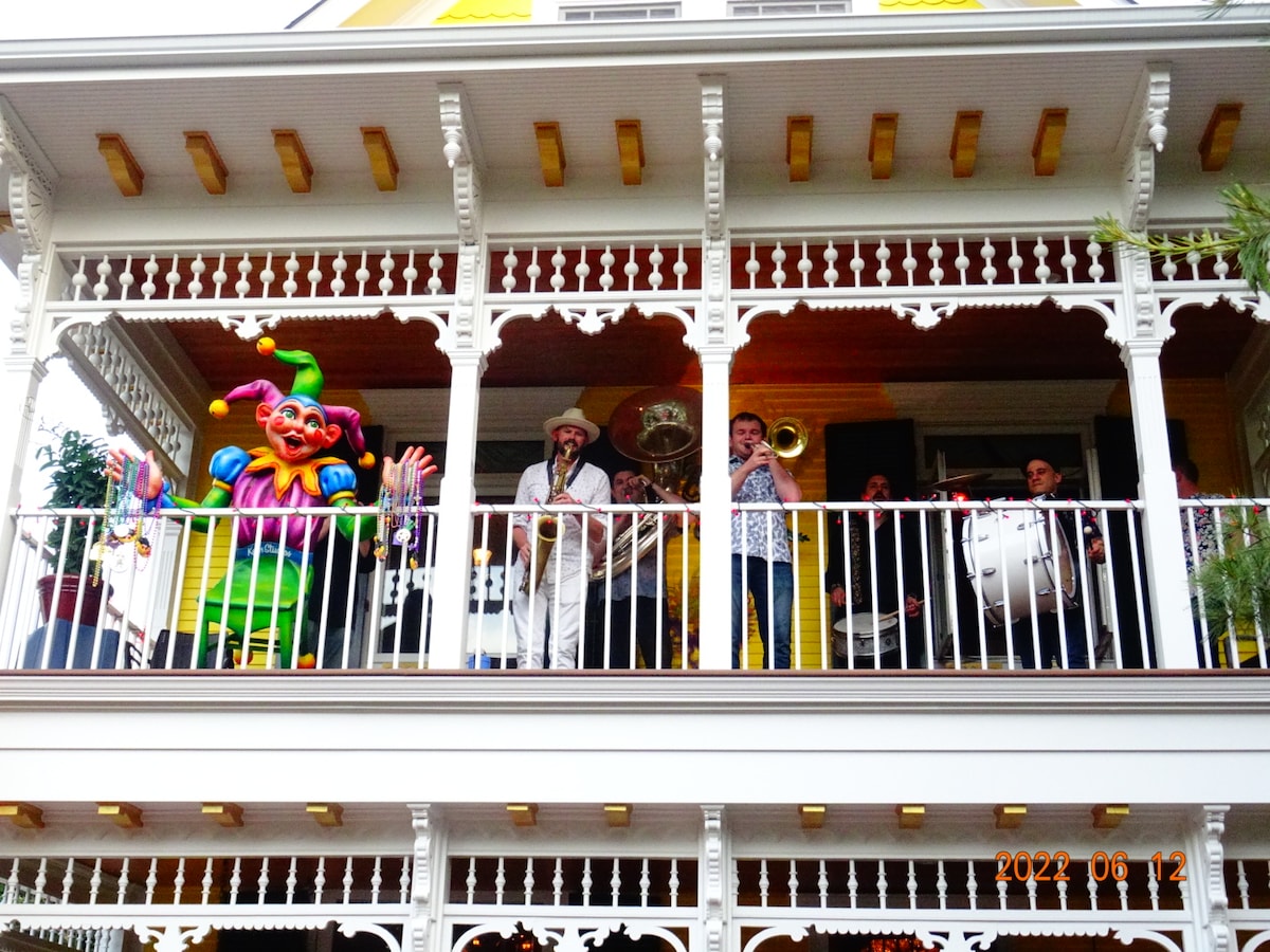 A vibrant balcony features a lively group of musicians performing. A jester costume adds a playful touch to the atmosphere. The intricate wooden railing and architectural details showcase the historic charm of the Mardi Gras House, while greenery enhances the inviting setting.