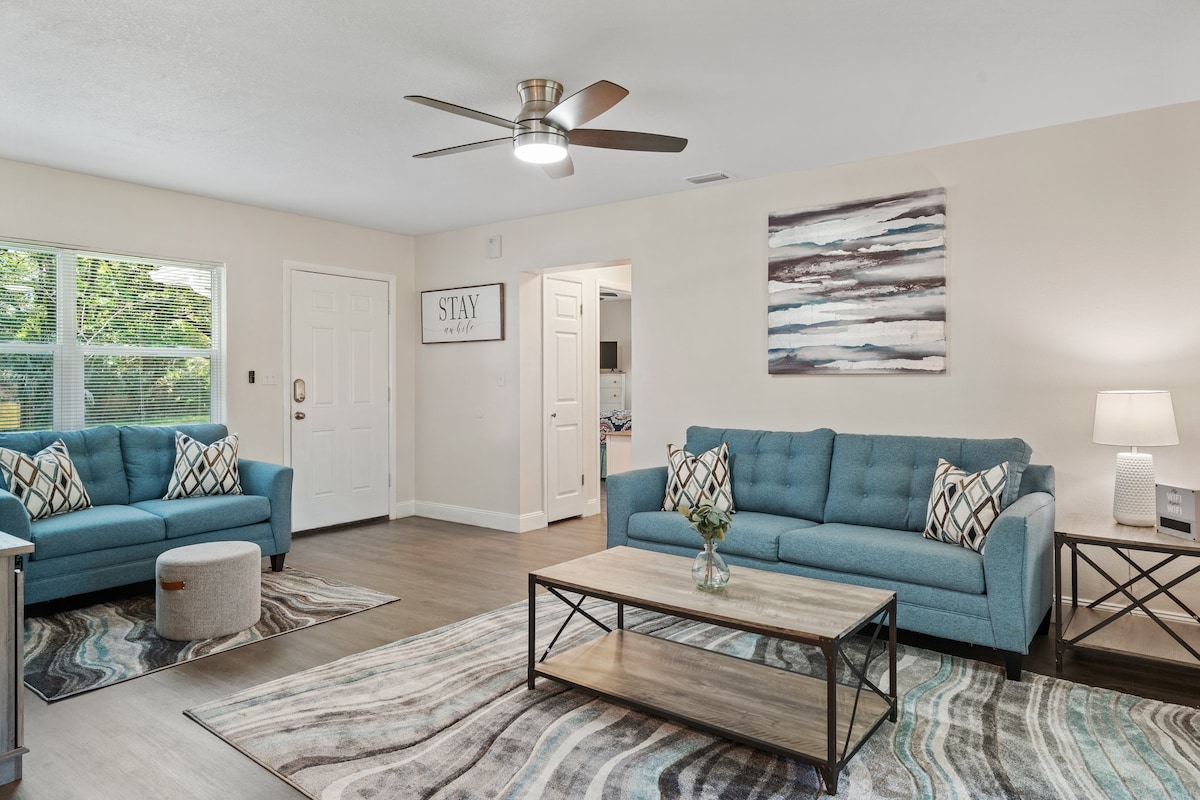 A light-filled living area features two soft blue sofas arranged around a coffee table. Neutral rugs anchor the seating, while a modern ceiling fan provides airflow. A wall art piece in soothing tones complements the space, with a welcoming entryway visible in the background.