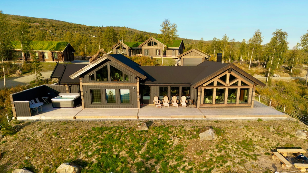 The mountain cabin features a spacious terrace with seating arrangements and a jacuzzi. Large windows provide views of the surrounding greenery. The exterior showcases wooden architecture complemented by a dark roof, blending harmoniously with the natural landscape.