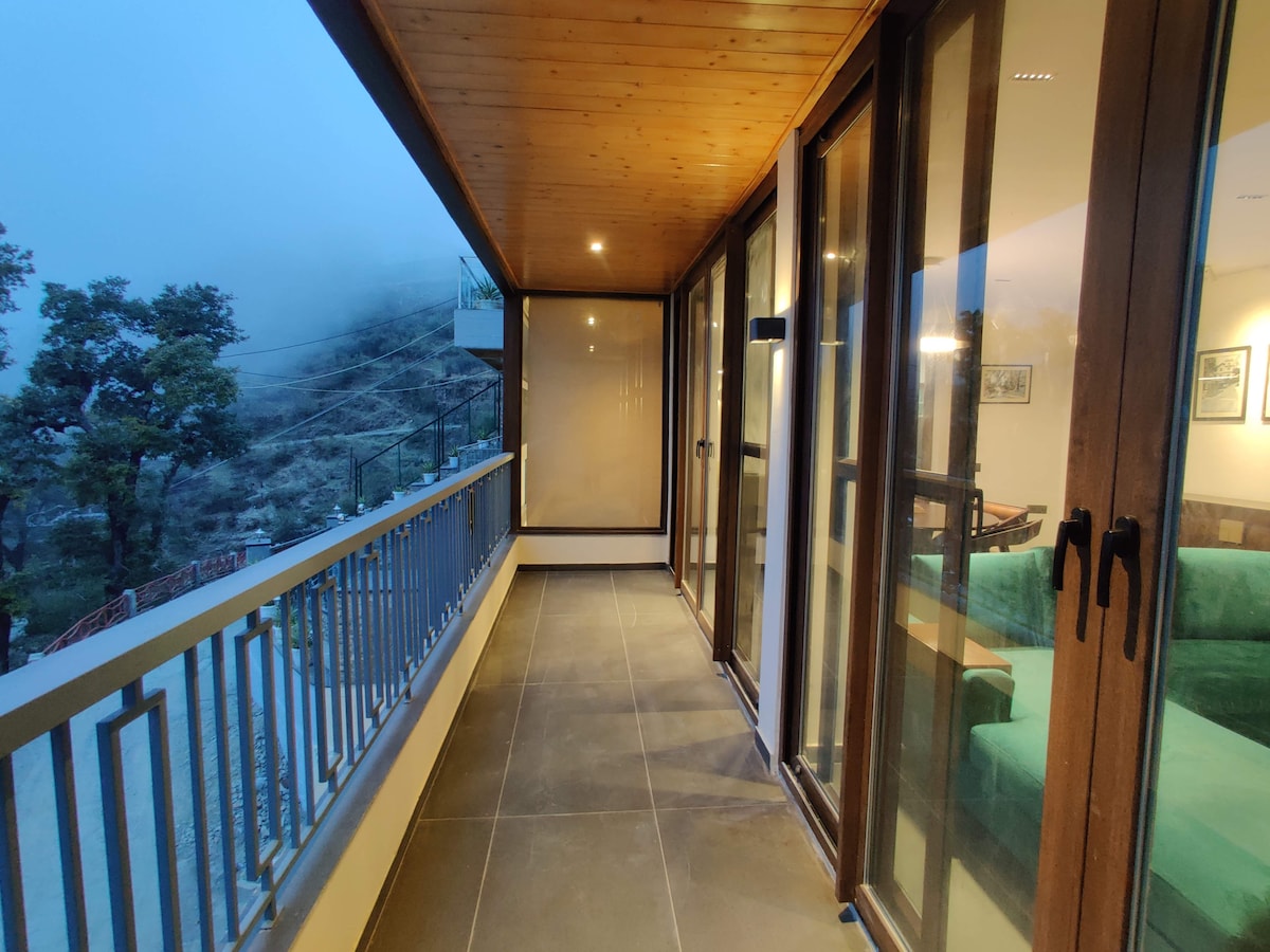 A balcony with large glass doors provides an unobstructed view of the surrounding landscape. The wooden ceiling complements the contemporary flooring. Soft lighting creates a serene ambience, inviting guests to enjoy the fresh air and tranquility of the hills.