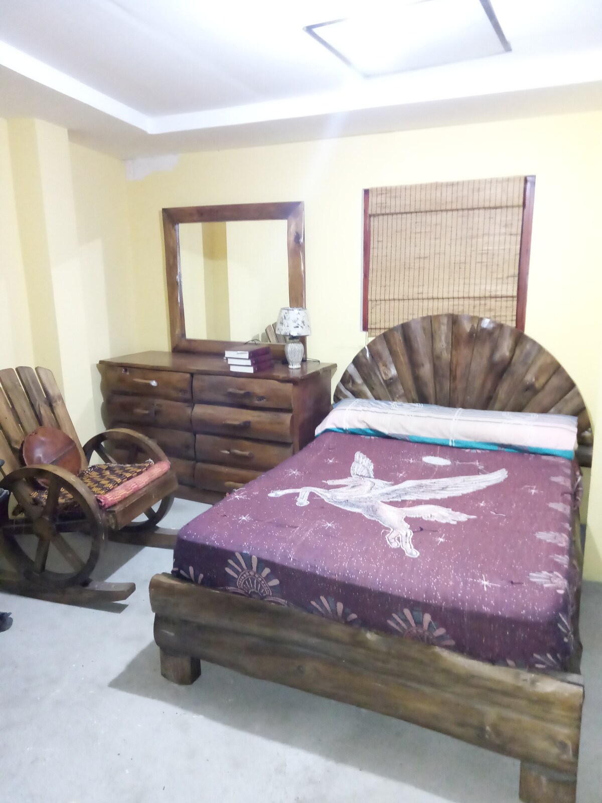 A warm room features a spacious bed adorned with a patterned blanket. A wooden dresser and a mirror are positioned nearby, enhancing the rustic feel. A unique wooden chair adds character, while soft lighting creates a cozy environment.