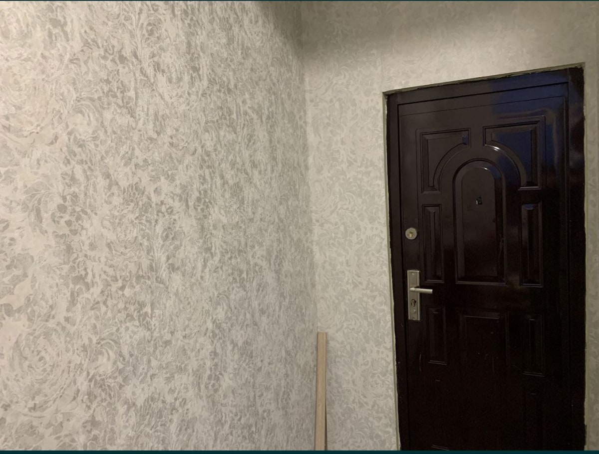 A simple entryway features light-colored patterned wallpaper, creating a soothing environment. A dark wood door with a metallic handle is centered in the image, providing access to the space. A wooden post stands on the floor, adding a touch of minimalism.