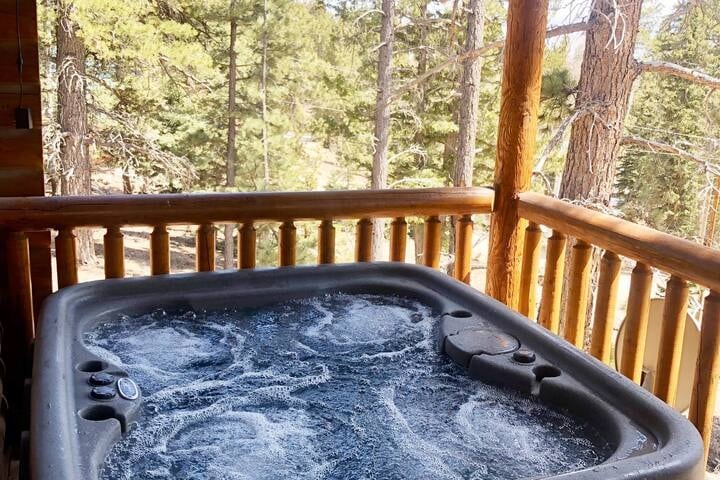 A hot tub sits on the wooden deck, surrounded by tall trees. Bubbles create a relaxing ambiance in the water, inviting relaxation while enjoying the surrounding natural landscape.