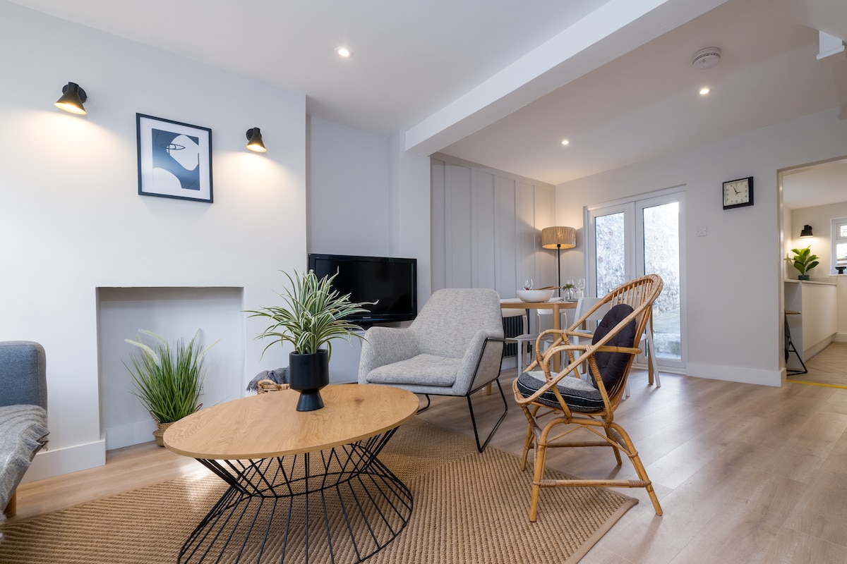 A light-filled living room features minimalist furnishings, including a round wooden coffee table and two armchairs in soft tones. A flat-screen TV is positioned against a wall, while double doors lead to a bright outdoor space. Textures are blended to enhance the calming atmosphere.