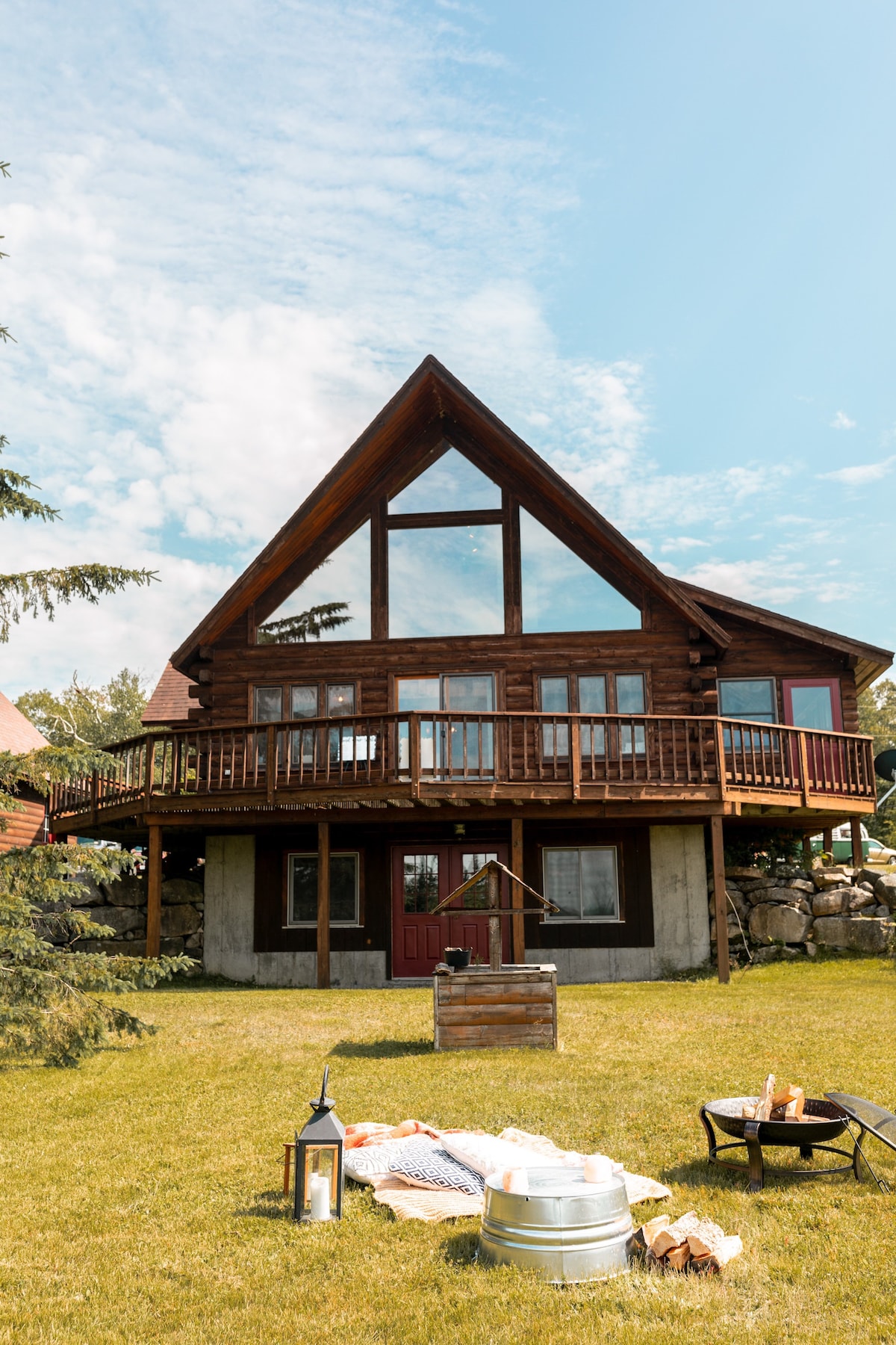 The A-frame log cabin features a striking triangular roof and large windows that allow natural light to flood the interior. A spacious wraparound deck provides outdoor seating options, while a neatly maintained lawn and a fire pit area enhance the outdoor appeal.